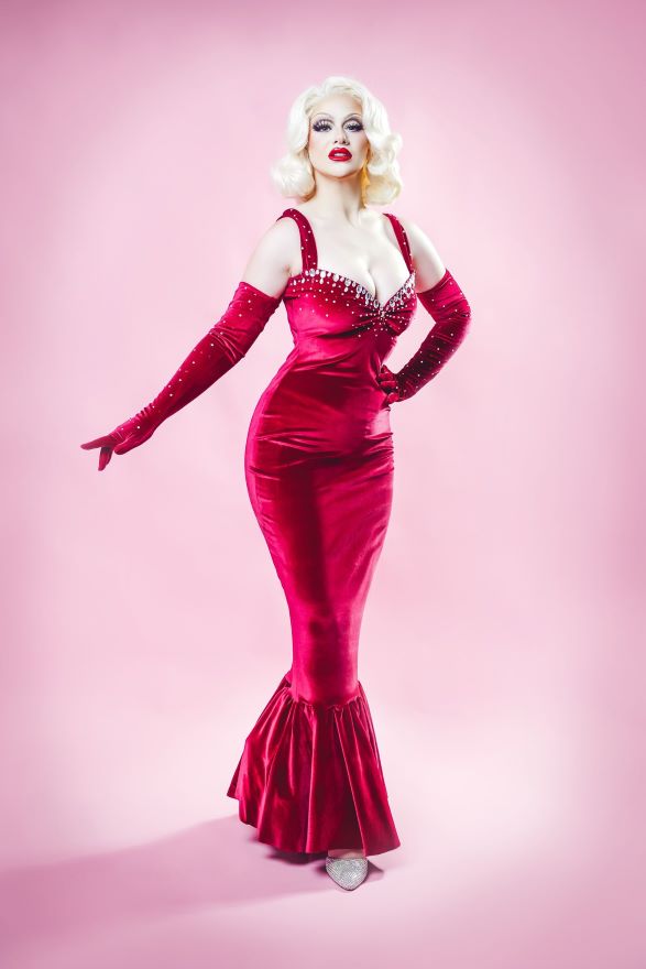 Pictured is Cece Desist - A Marilyn Munroe-style performer with blonde wig, red dress and gloves, in front of a pink backdrop