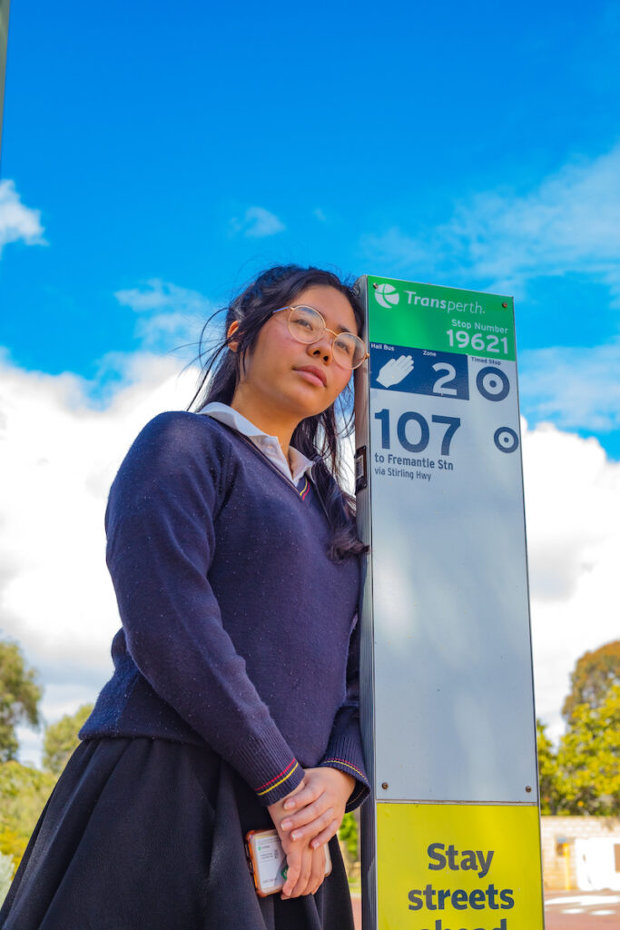 An image of a young asian woman in a school uniform leaning against the 107 bus route stand. 