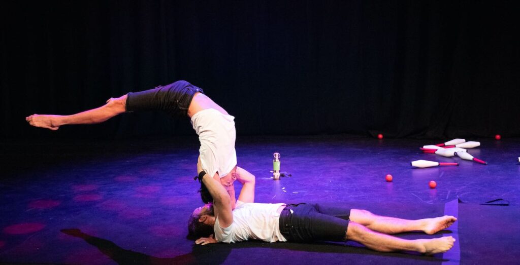 Performers from Acrobatch Circus. Pictured one man lies on the floor, holding the arms of another man who is in a handstand that arches over at a seemingly impossible angle.
