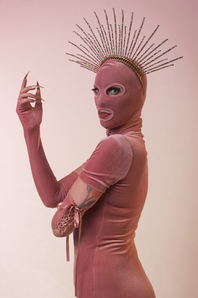 Woman in pink, velour dress, long gloves and a balaclava stands in front of a pink gradient backdrop, wearing a sparkling crown.