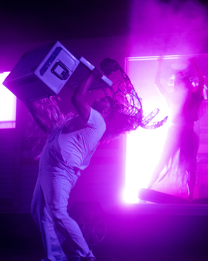 An actor from Fremantle Theatre Company dressed in white t-shirt and overalls tips an esky of ice over himself. The scene is lit in a purple glow