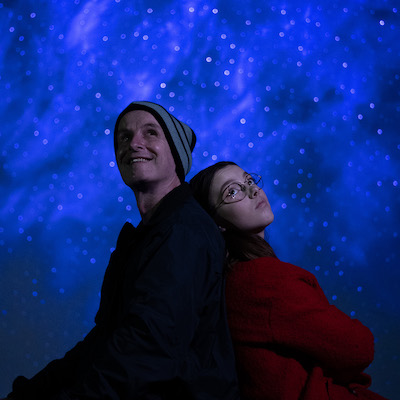 A promotional image from the Fringe World show 'Freeze'. Pictured is a male and female leaning back to back, both look into the distance. 