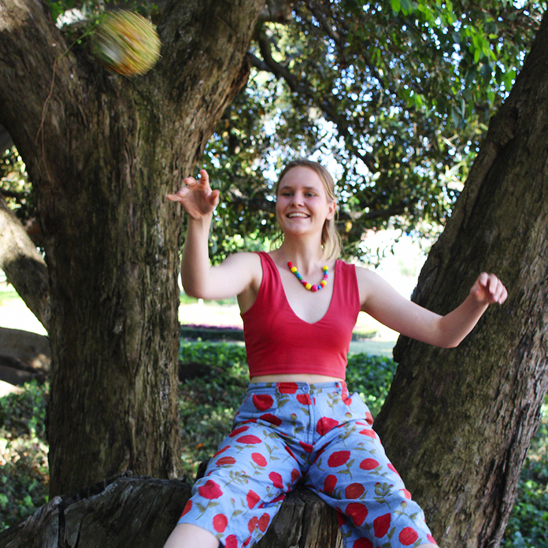 Promotional image from She's Terribly Greedy. Pictured: A woman sitting on a tree. She wears a red singlet and blue pants with red roses. She holds a happy expression.