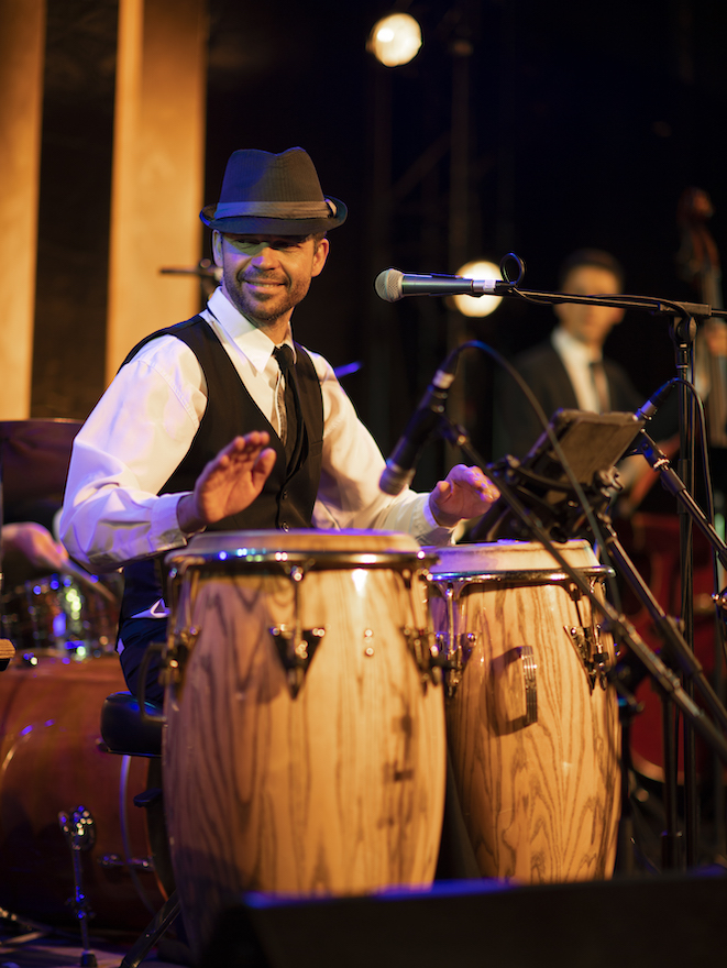 An image of a man from Havana Nights playing a set of the bongo drums with a microphone towards his face. He is dressed smartly in a white long-sleeve business shirt, black tie, black vest and fedora hat.