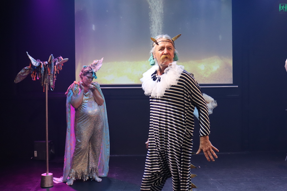A snapshot from Conversations with a Fish. Pictured a man stands, poised, his arms slightly behind his torso, his eyes focused to something beyond the frame. He wears a black and white striped onesie and horns.