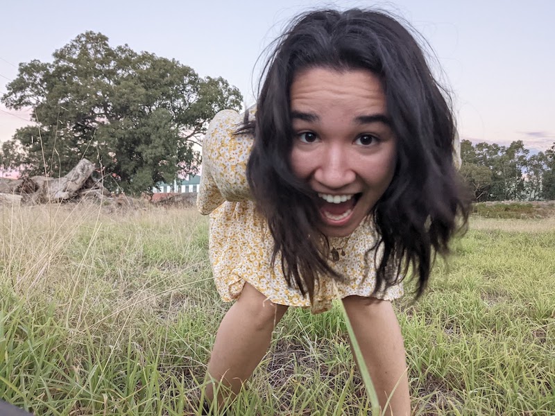 107 writer and composer Michelle Gould. Pictured is a young asian woman standing in a field. She's leaning over and smiling wildly at the camera.