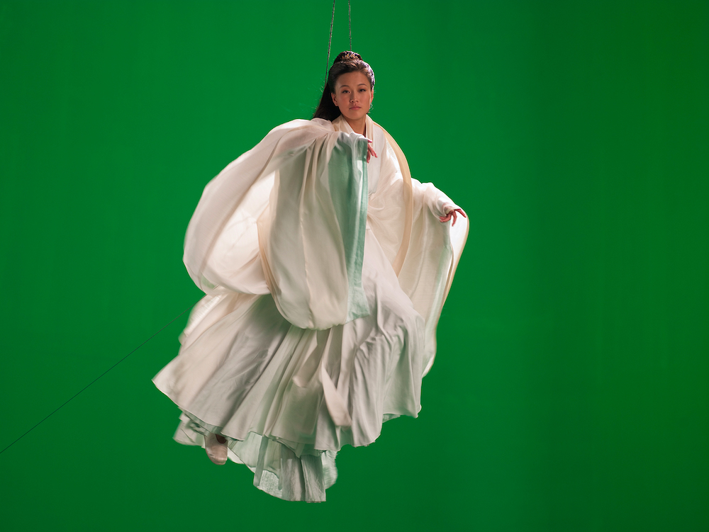 A photo of a young Asian woman, suspended in the air by wires, dressed in billowing white fabric, against a green screen.