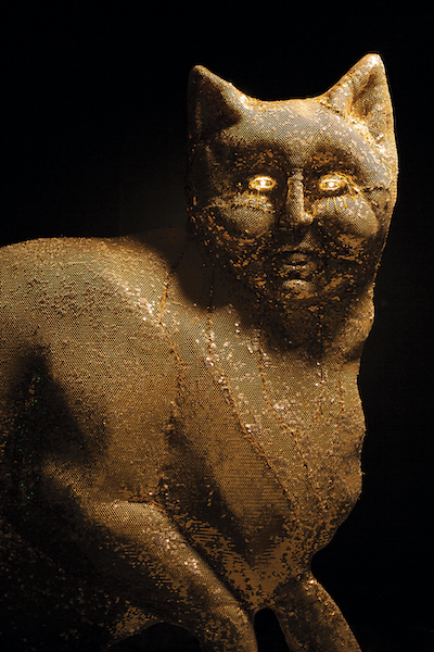 A sculpture of a cat, from the Wardan visual arts exhibition, with a human-like face. The cat is textured in a bronze/gold material and its eyes glow golden.