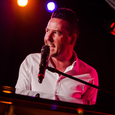 A promotional image from the Fringe World show 'Michael Griffiths: Greatest Hits'. Pictured is a close-up of a man singing into a microphone behind a piano. 