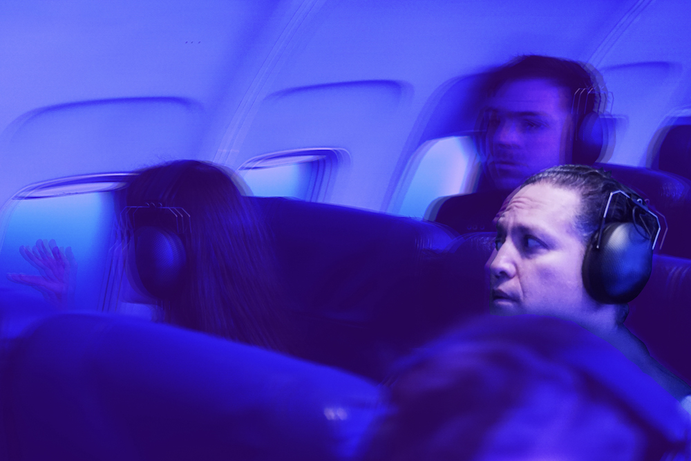 An image from Fringe World's 2022 sensory experience FLIGHT of the articles review of FLIGHT and COMA. Pictured is three individuals sitting in airplane seats. The plane is flooded with blue, and each person has a bulky set of headphones on and have a distressed look on their faces.