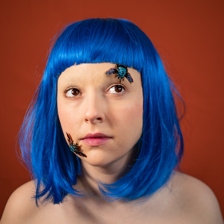 A headshot of a young woman with bright blue hair cut in a long bob with a blunt fringe. On her face are bright blue winged beetles. She looks up to one corner.