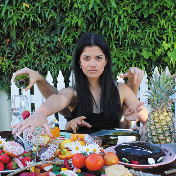 A promotional image from She's Terribly Greedy part of the Summer Nights curation. Pictured is a woman with dark hair sitting at a table of food. From behind two sets of hand appear, one set pouring a drink the other reaching forward.
