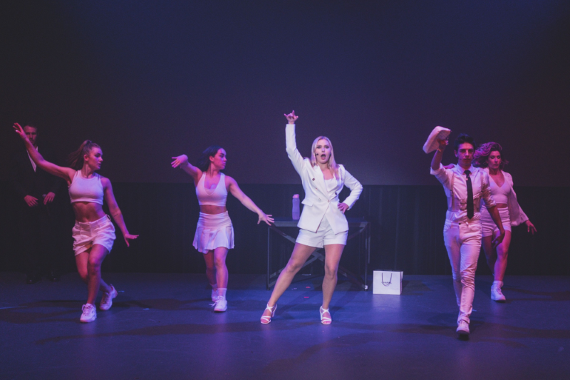 Promotional image from Sugar & Ice, oictured is 5 individuals all in white mid-dance. In the spotlight a blonde woman stands one foot pointed as she sings.