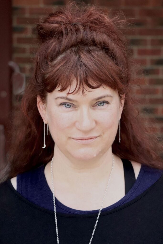 Pictured is a headshot of Teresa Izzard, the director and producer of 'This Is Where We Live' for Fringe World 2022. Izzard is a woman with red hair wearing a black shirt with a blue singlet underneath, standing against a red brick wall.