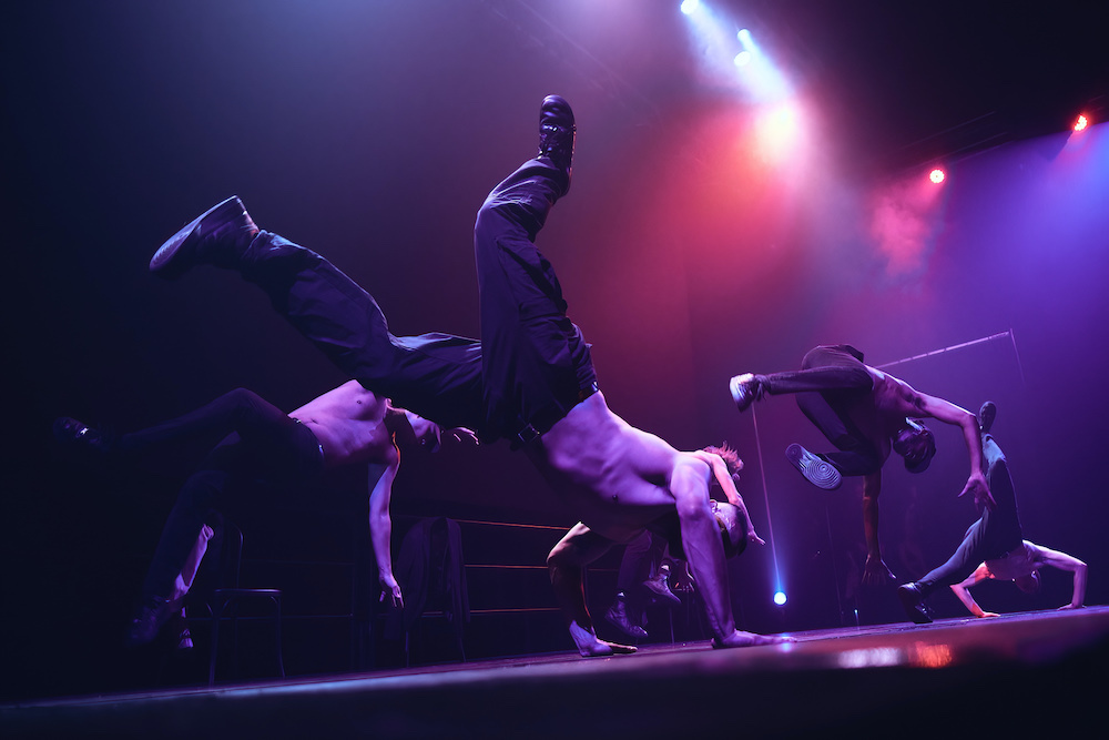 Four male dancers from 'The Underground' performance. Pictured are these men on stage in the middle of acrobatic break dance moves. The stage is dark and lit with pink spots. The men wear black pants and shoes but no shirts.