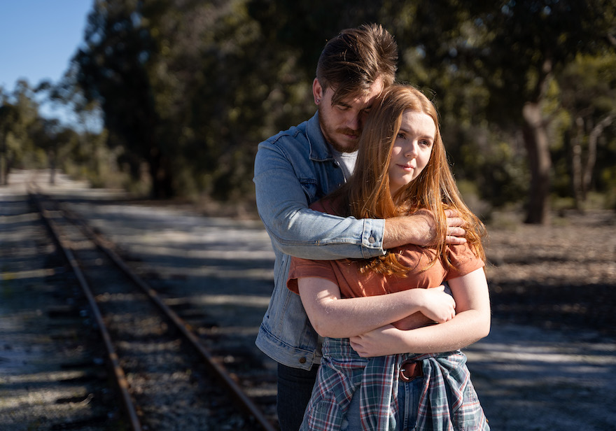 This Is Where We Live promotional shot. Pictured is a male and a female. The male stands behind the female his arms wrapped around her shoulders and torso, whilst the female stands looking into the distance holding the mans hand around her torso.