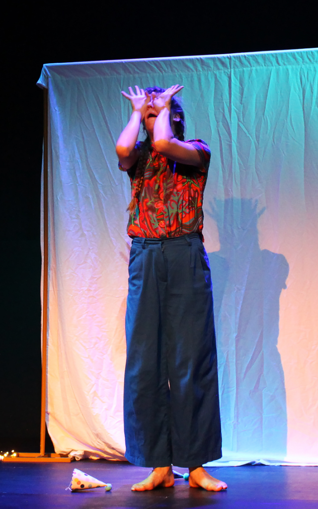 A performer from Dreams of a Lonely Planet in a red shirt and long baggy pants stands with hands over her face