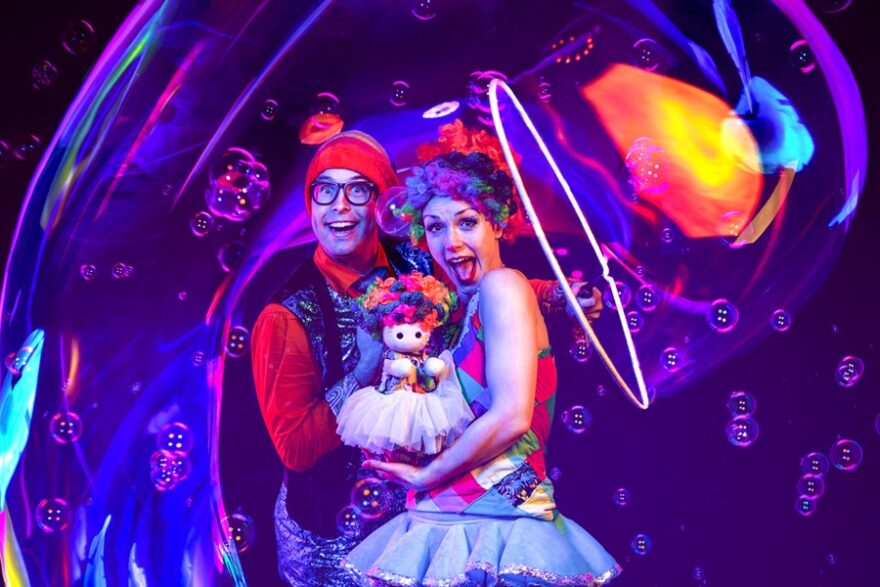 Two people stand inside an enormous bubble, holding a puppet