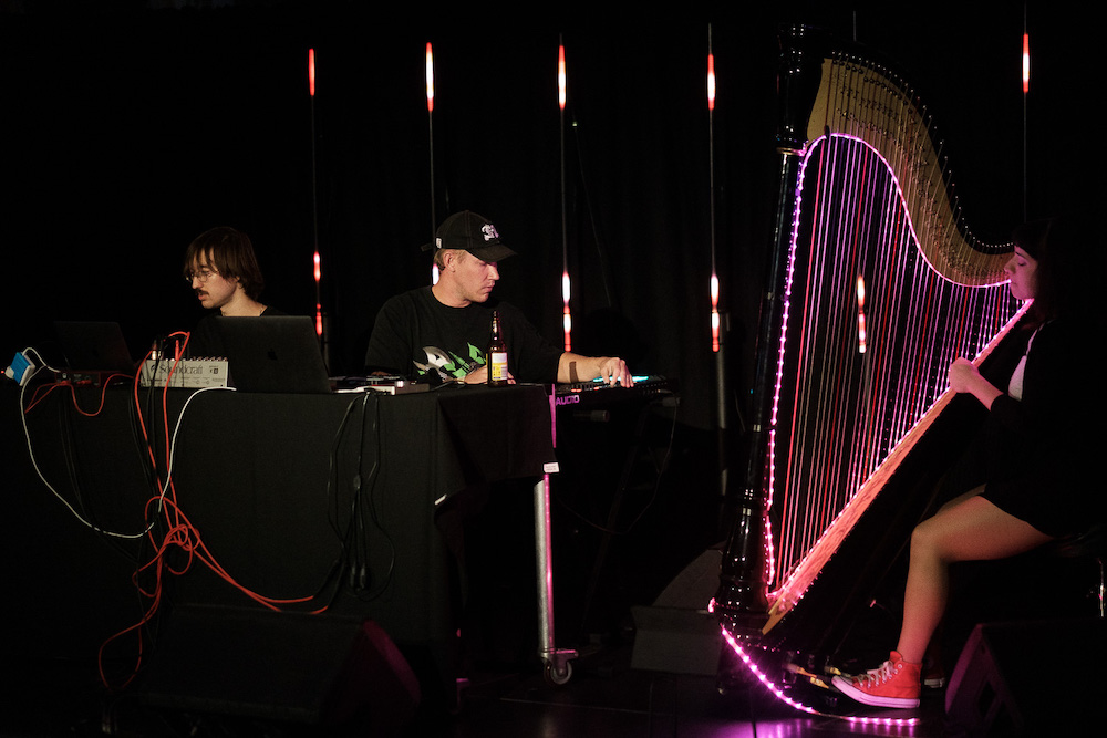 Two men sit at a table with laptops. next to them is a woman playing a floor harp with its strings illuminated in pink