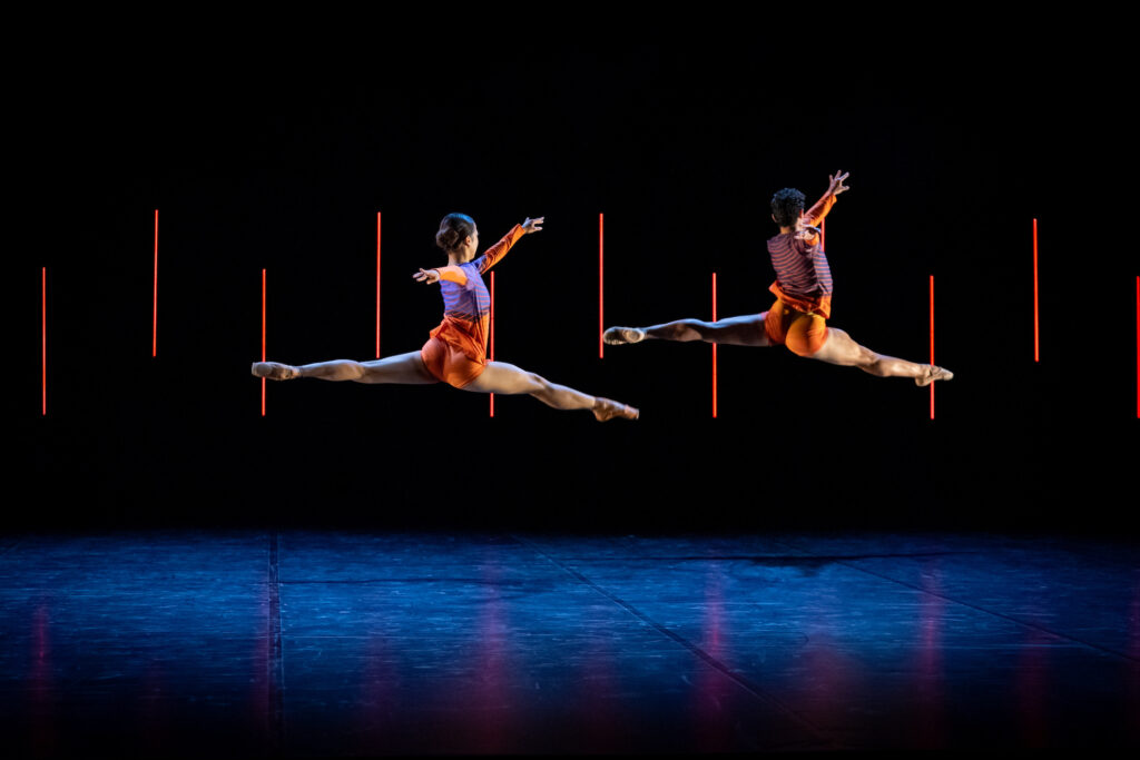 Two ballet dancers on stage, leaping into the air with legs and arms at full stretch