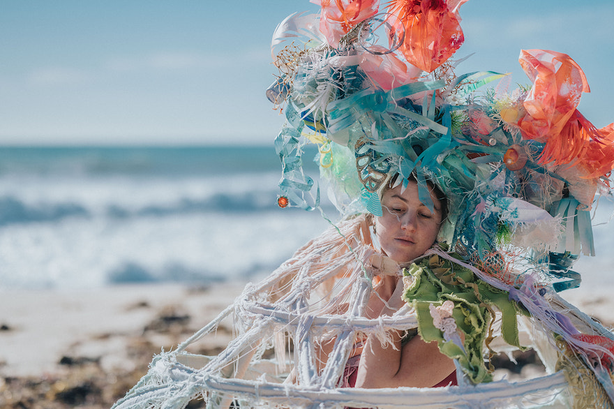 An image of Talitha Maslin in costume for Siren. Pictured is a woman on the beach wearing a large headpiece that looks like coral and jellyfish.