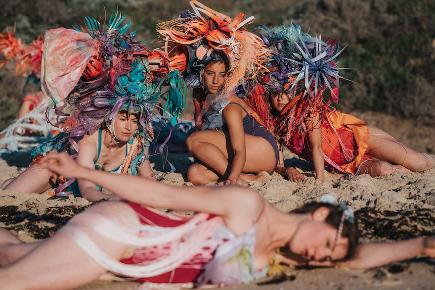 A promotional image from Talitha Muslin and Gogi Dance Collective show Siren. Pictured is three women in the background wearing large orange and blue headpieces overlook another dancer who lies down in the foreground.