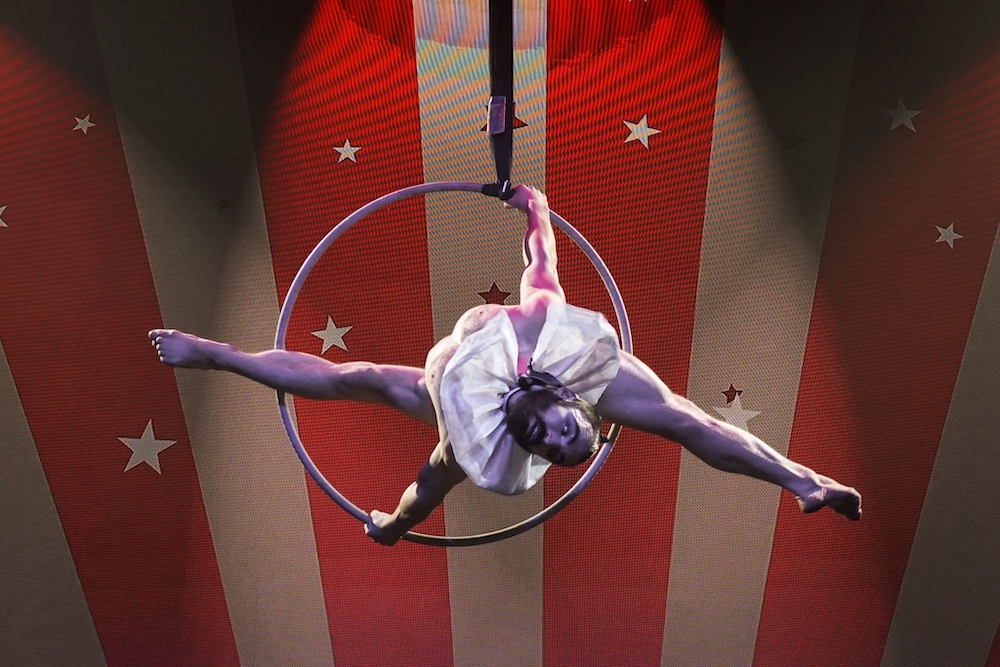 An image from BarbieQ and Kinetica's show 'P!nk: The Circus'. Pictured is a male suspended in the air male hoop aerialist holds the hoop with both hands while his legs split above the hoop.