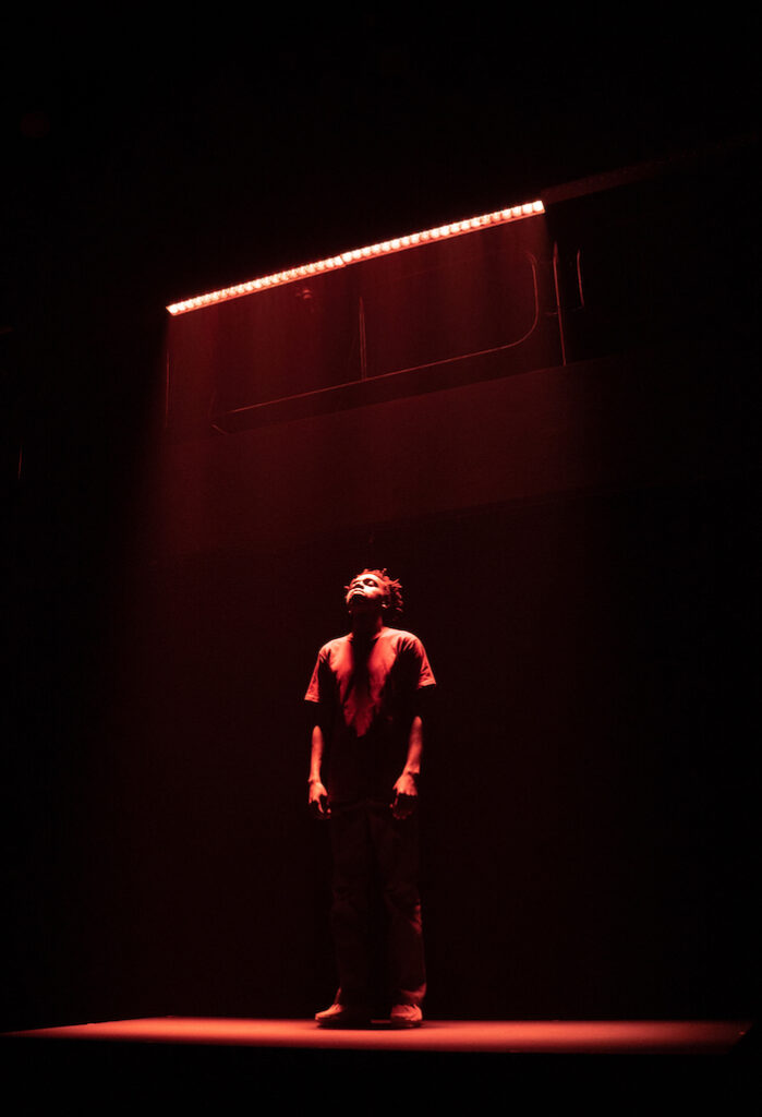 An image from Oliver Twist's show JALI, pictured a man stands looking up to the sky a red light engulfing him.