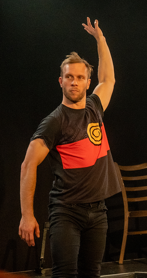 Pictured is Joel Bray, performing in his show I Liked It But. Bray wears an indigenous flag t-shirt. He stands in an el matador-esque pose.