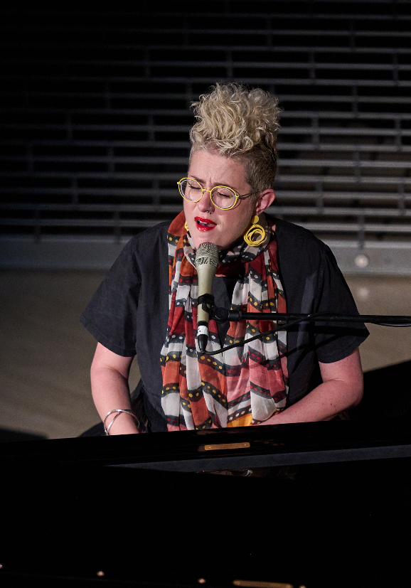 Singer Katie Noonan sits behind a keyboard as she sings into a mic. The woman wears a black t-shirt, red and white scarf and a yellow pair of glasses.