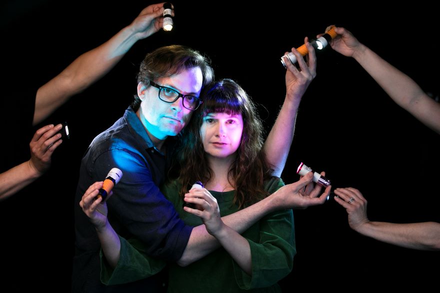 A promotional image from Stage Changers. Pictured a man and woman in a dark room are illuminated by hands reaching toward them with torches