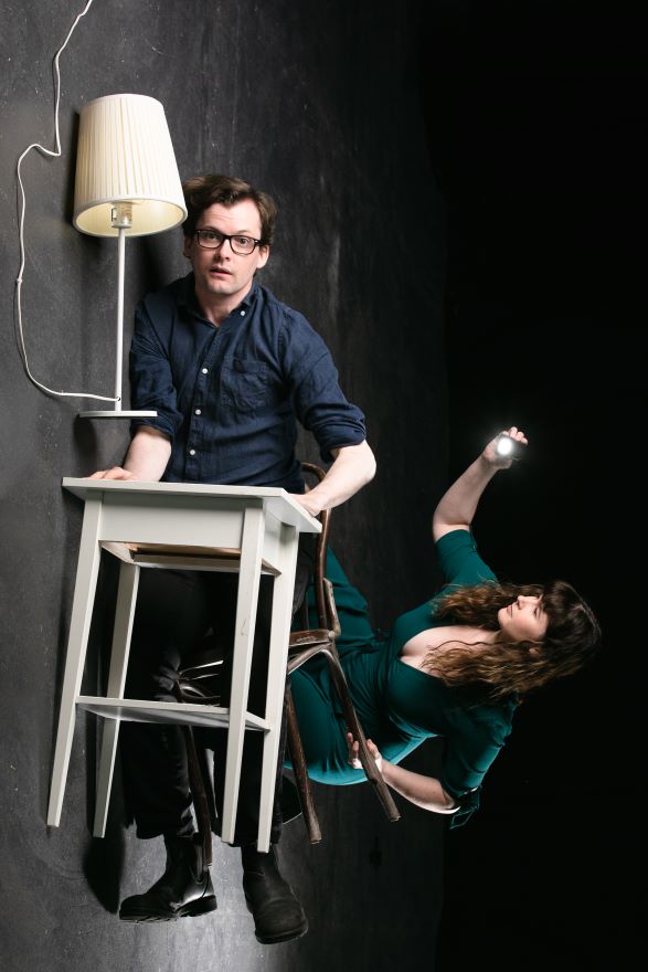 A promotional image from Stage Changers. Pictured a man is drifting upward from the chair, desk and lamp that seem to float away from him. His dangling feet defy gravity, as does a woman who is drifting at right angles to him, shining a torchlight.  