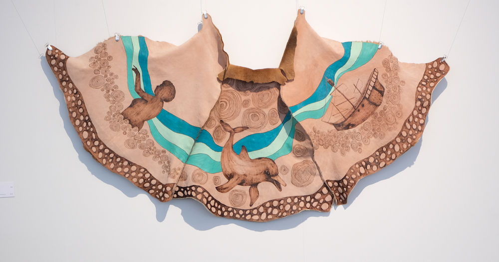 Artwork from the Strangers on the Shore exhibition, picture is a kangaroo skin decorated with an image of a person, dolphin and ship.