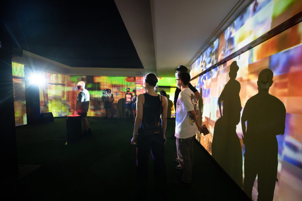 An image from Ta-Ku and friends Songs to Experience, pictured are group of people standing in a room lit up by a projector.