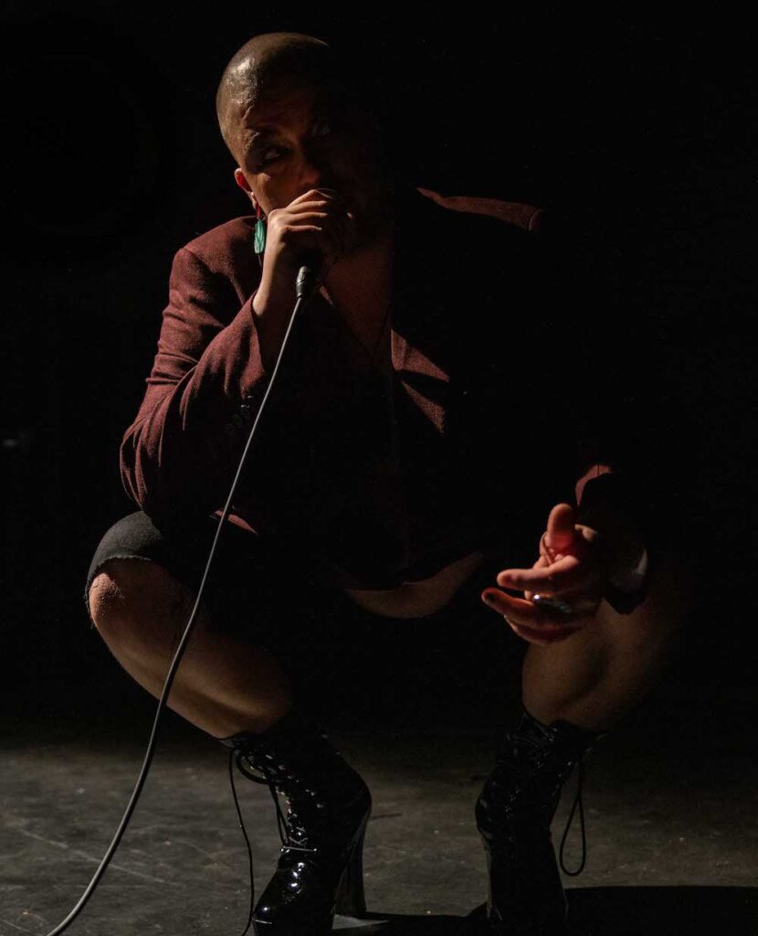 Takatāpui creator Daley Rangi is pictured squatting holding a microphone to his face. The room is dark and Rangi is half coverd by the shadows as they look directly at the camera.