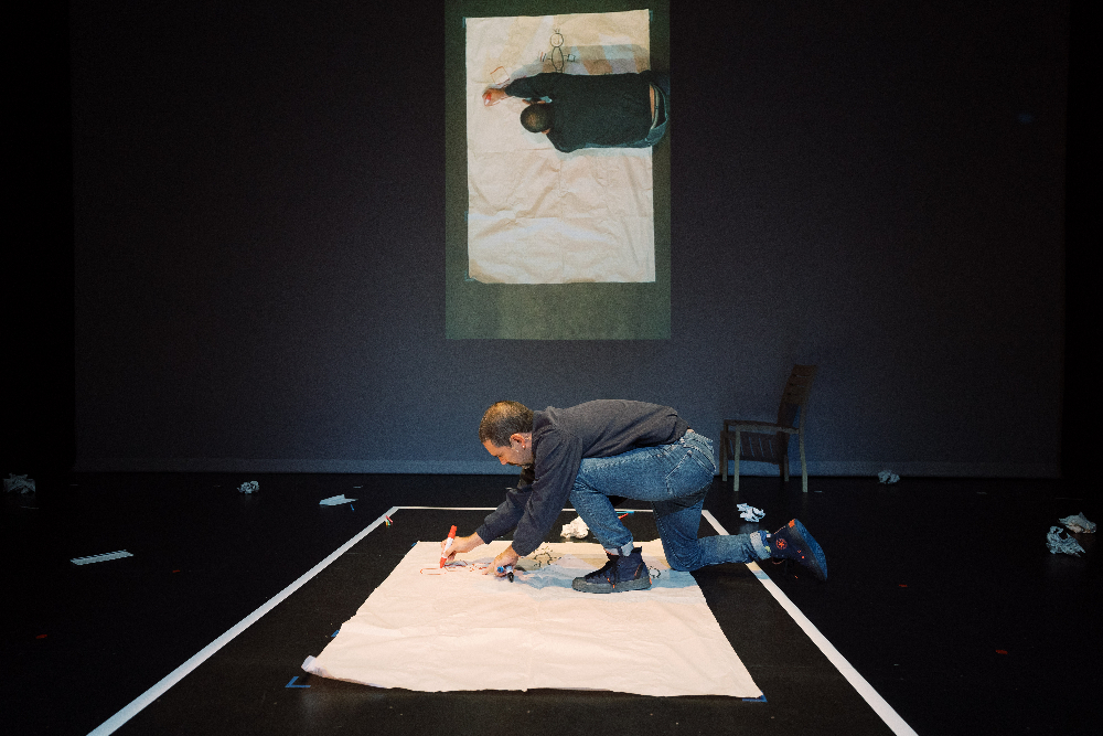 A man kneels on a stage as he draws on a large sheet of paper with a coloured marker