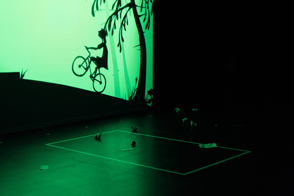 At the back of an empty stage is a stylised silhouette of a tree and a a child riding a bicycle up a slope. The child is doing a wheelie. The sky is luminous green.