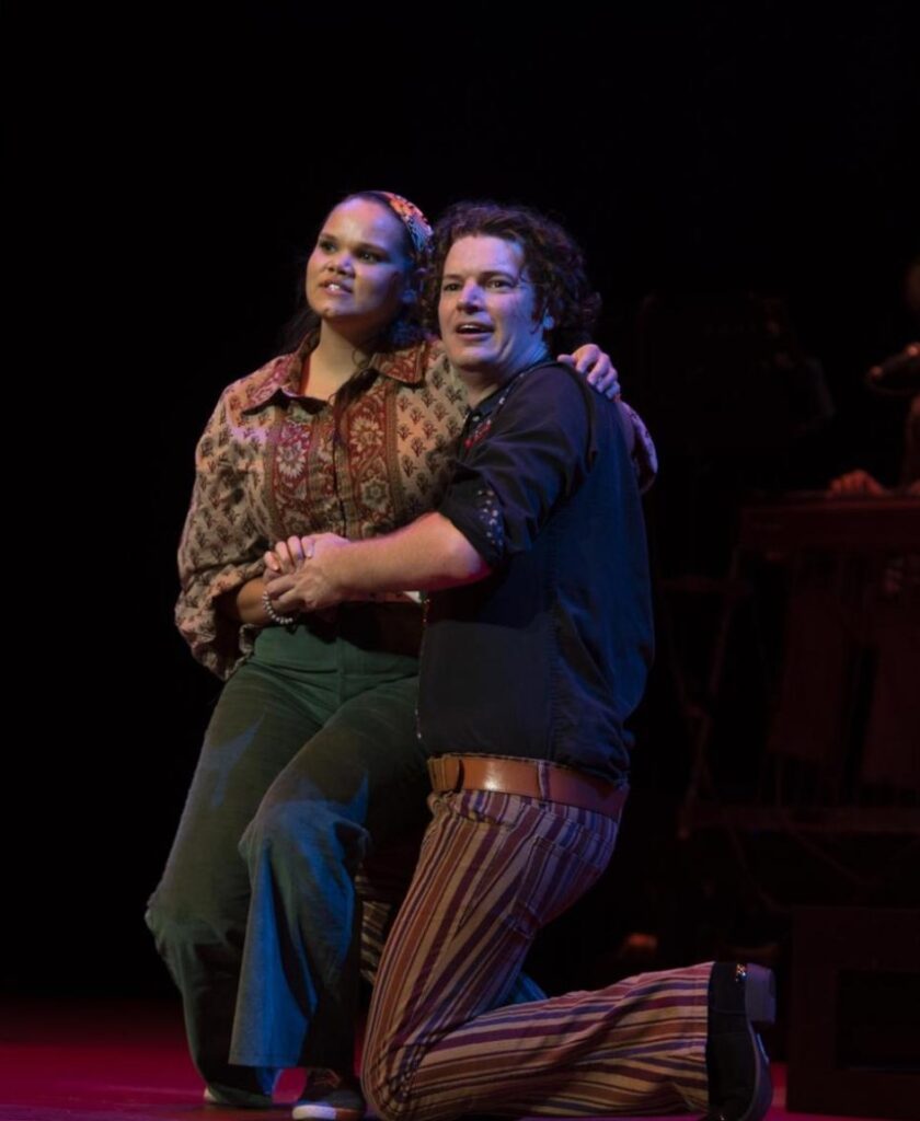 An image from Yirra Yaakin Theatre Company's performance of Panawathi Girl, pictured a woman in a blouse and pants sits on a man who is kneeling. They both look out into the distance.