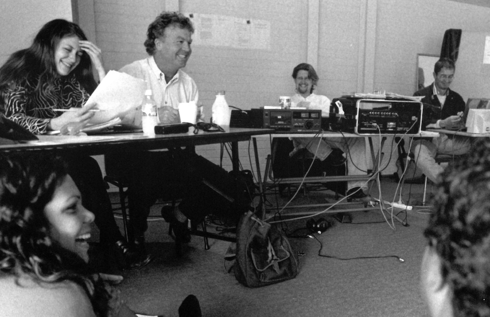 A black and white photo of a Andrew Ross AM and group of people sitting around desks. Everyone is smiling, as though at a joke. There's sound equipment on one of the desks.