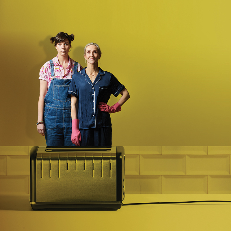 Two women stand in a yellow room. In front of them is a chrome toaster.