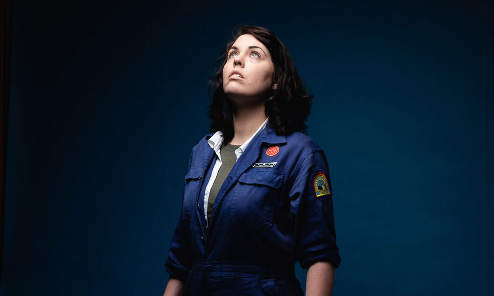 A woman dressed in a navy boiler suit, decorated with woven badges, looks up at something out of shot.