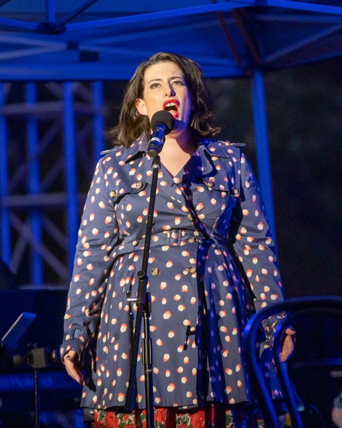 A FFO performer for the Opera La Boheme. Pictured a woman in a dark coat with cream flecks on it sings into a microphone
