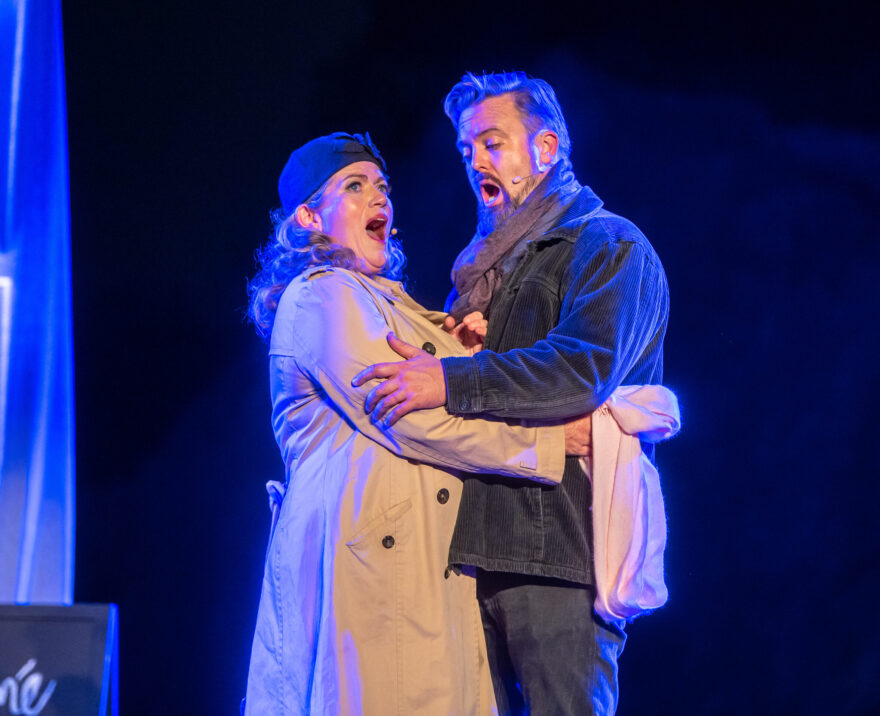 Two character from FFO perform on stage together. They are a man and woman dressed in coats and scarves hold each other's arms as they sing on a blue lit dark stage
