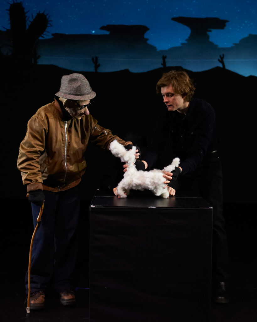 It's Dark Outside character of an old man from the Last Great Hunt's show leans on a walking stick and pats a puppet that looks like a small white dog, being held by a man wearing all black