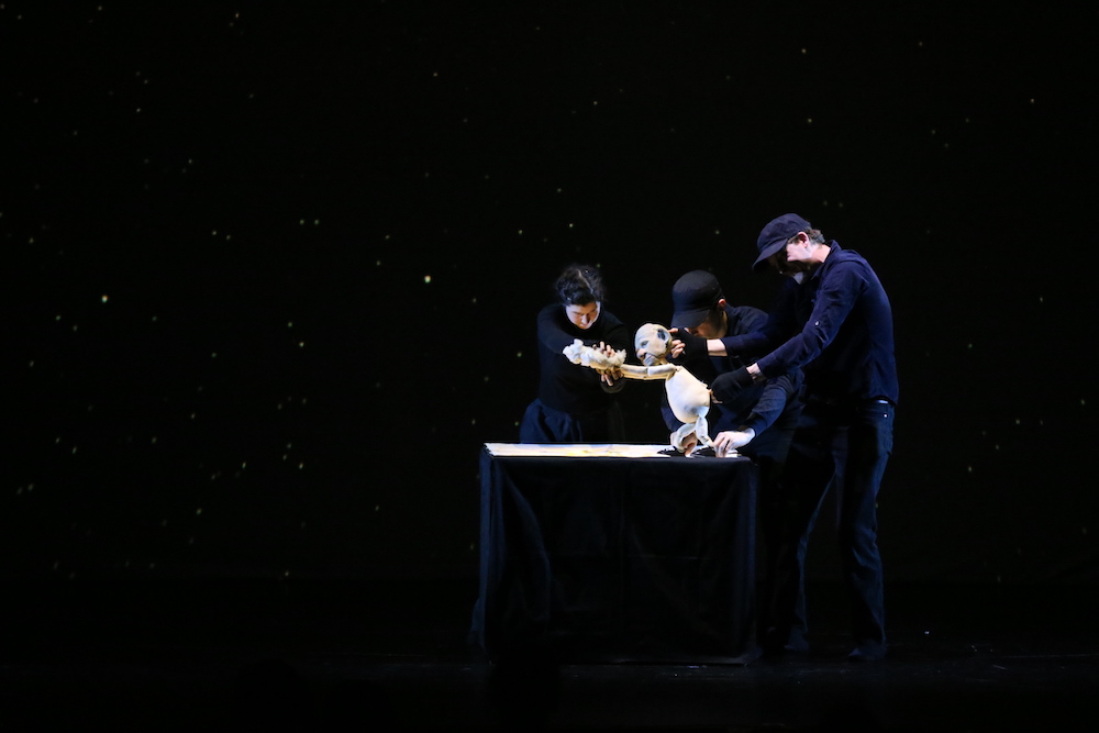An image from the performance of It's Dark Outside, pictured is three actors in black manipulating a puppet against a back drop filled with stars.