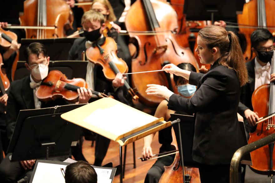 Jen Winley conducting WASO, she wears a black suit with a pony tail. The musicians wear masks.