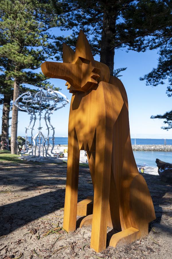 A sculpture of a dog, in steel. It sits on scraggy grass under pine trees. We can see a beach in the background.