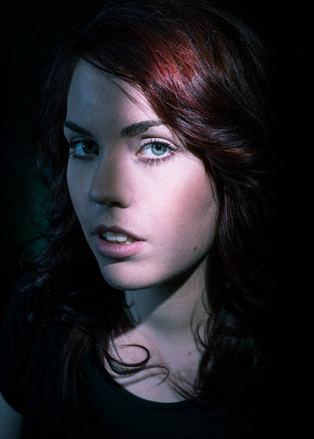A headshot of a woman with red hair and green eyes. Her head is turned slightly away from the camera, but her eyes are turned to the lens.