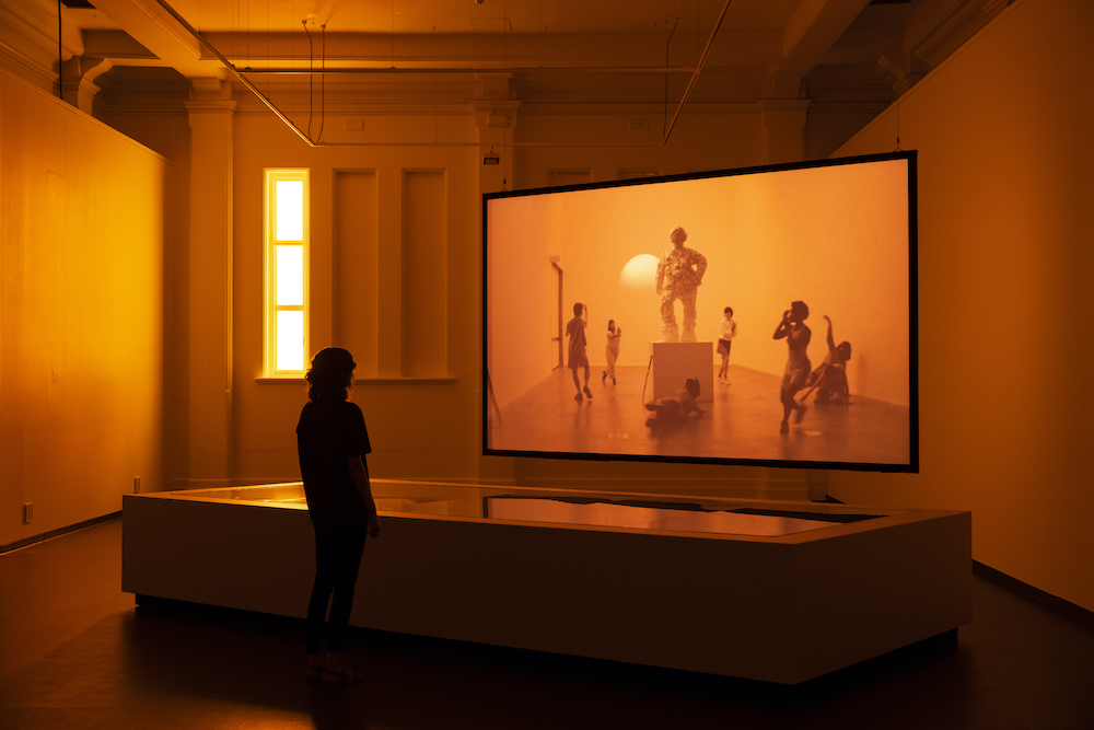 Pictured is the exhibition titled 'monumental' a person stands in a room drenched in orange light, they are watching a screen, on which we can see another orange light filled room, and dancers moving around a lumpy statue of a person.