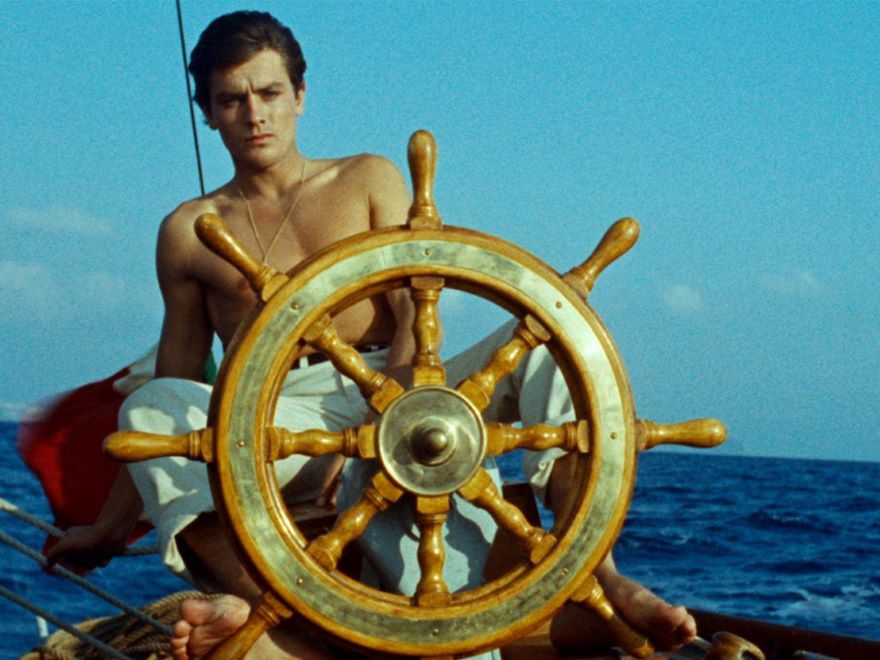 A vintage, pixelated photo of a young man wearing no shirt and sitting behind the wooden tiller of a boat with blue sky and ocean behind him